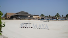 A sunlit beach area with silky white sand and a resort-style restaurant in the background. The sandy expanse leads to various thatched umbrellas and lounge chairs arranged for sunbathing. Palm trees are visible around the area, and a sign displays the text '#ALMAYA' near the forefront.
