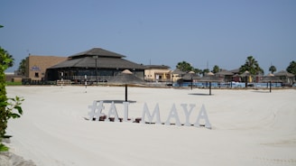 A sunlit beach area with silky white sand and a resort-style restaurant in the background. The sandy expanse leads to various thatched umbrellas and lounge chairs arranged for sunbathing. Palm trees are visible around the area, and a sign displays the text '#ALMAYA' near the forefront.