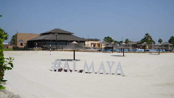 A sunlit beach area with silky white sand and a resort-style restaurant in the background. The sandy expanse leads to various thatched umbrellas and lounge chairs arranged for sunbathing. Palm trees are visible around the area, and a sign displays the text '#ALMAYA' near the forefront.