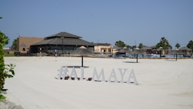A sunlit beach area with silky white sand and a resort-style restaurant in the background. The sandy expanse leads to various thatched umbrellas and lounge chairs arranged for sunbathing. Palm trees are visible around the area, and a sign displays the text '#ALMAYA' near the forefront.