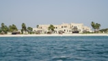 A luxurious beachfront property with a large, elegant building set behind a pristine sandy beach. Palm trees and small thatched structures provide shade along the shoreline. The sea in the foreground is calm and blue, contrasting with the light-colored architecture of the estate.