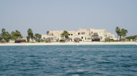 A luxurious beachfront property with a large, elegant building set behind a pristine sandy beach. Palm trees and small thatched structures provide shade along the shoreline. The sea in the foreground is calm and blue, contrasting with the light-colored architecture of the estate.