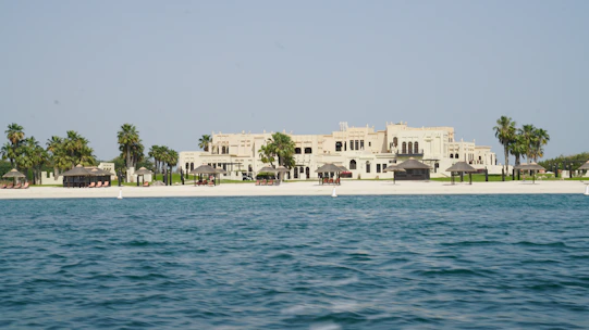 A luxurious beachfront property with a large, elegant building set behind a pristine sandy beach. Palm trees and small thatched structures provide shade along the shoreline. The sea in the foreground is calm and blue, contrasting with the light-colored architecture of the estate.