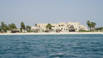 A luxurious beachfront property with a large, elegant building set behind a pristine sandy beach. Palm trees and small thatched structures provide shade along the shoreline. The sea in the foreground is calm and blue, contrasting with the light-colored architecture of the estate.