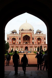 A large, ornate building with Mughal architecture is framed by a dark archway. The structure features a prominent central dome and two smaller domes on either side, made of white marble. The building's facade is adorned with red sandstone and white marble, showcasing intricate designs. People are walking towards the structure, and the surrounding area includes manicured gardens.