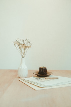 A minimalistic setup featuring a white vase with delicate dried flowers and a small dessert on a brown plate, positioned on a neat stack of papers on a wooden table. The background is a soft neutral color, enhancing the serene and simple composition.