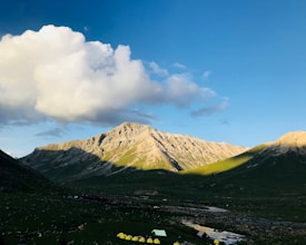 A scenic mountain landscape with a clear blue sky and large fluffy clouds casting shadows over the rugged peaks. In the foreground, a line of small yellow tents is set up on a grassy plain, suggesting a campsite. The sunlight highlights patches of green on the mountain slopes, creating a picturesque contrast.