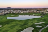 A serene landscape featuring a well-manicured golf course with sand traps and a large pond. In the background, there are rows of suburban houses and gently rolling hills under a soft, pastel-colored sky indicating either sunrise or sunset.