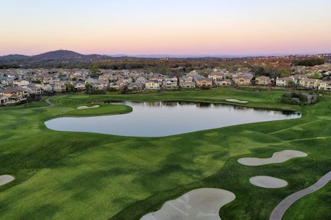 A serene landscape featuring a well-manicured golf course with sand traps and a large pond. In the background, there are rows of suburban houses and gently rolling hills under a soft, pastel-colored sky indicating either sunrise or sunset.