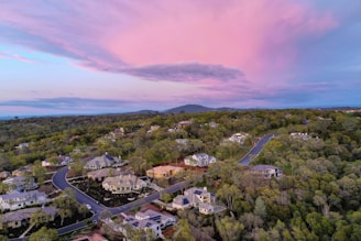 A serene suburban neighborhood with sprawling homes nestled amidst lush greenery. The winding roads connect the houses, surrounded by dense trees and foliage. Overhead, the sky is painted with a blend of soft pink and blue hues, indicating dawn or dusk. In the distance, gently rolling hills add depth to the landscape.