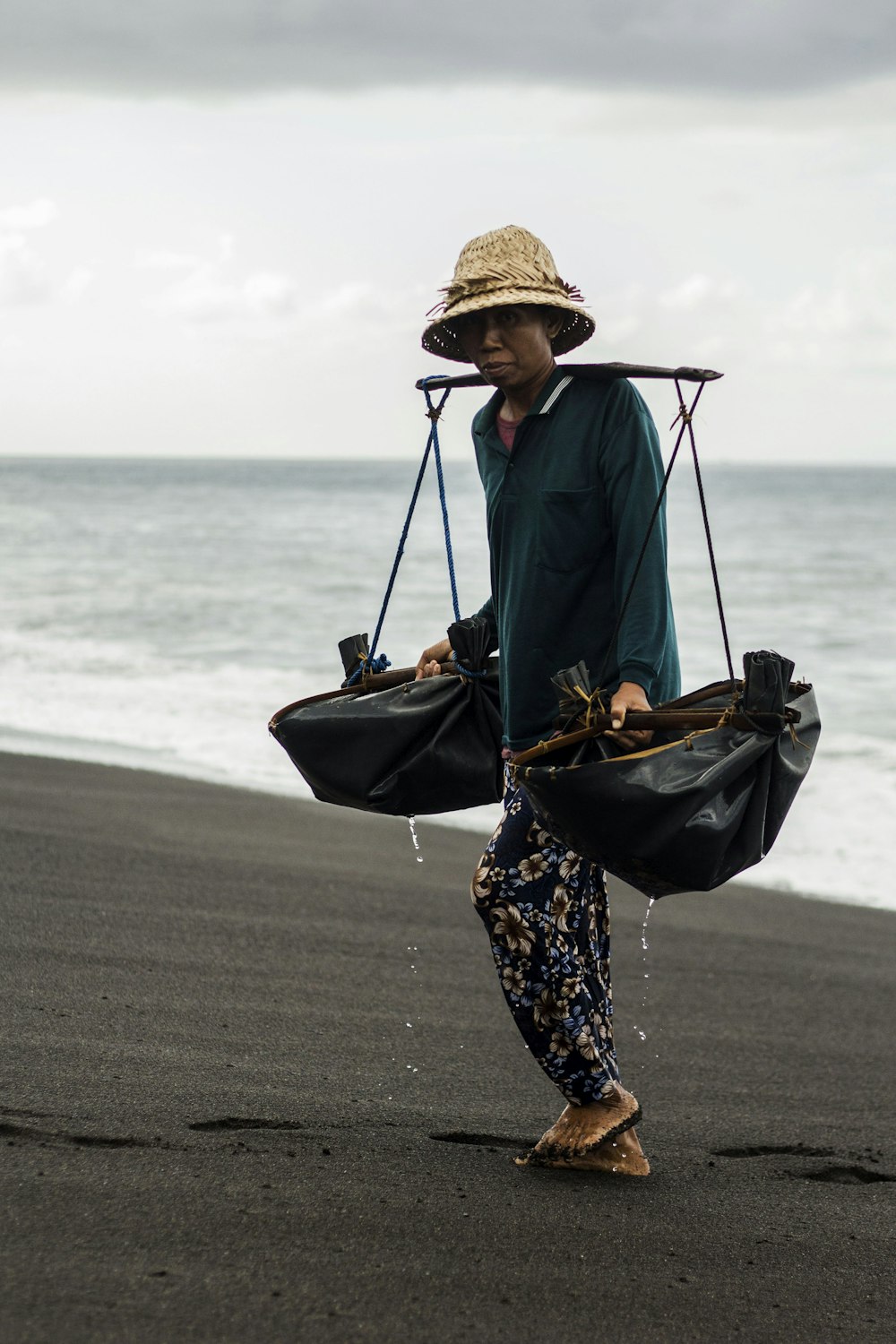 woman in blue jacket and black and white floral pants wearing brown hat standing on beach