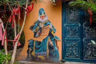 A mural of a monkey dressed in elaborate traditional Chinese attire with intricate designs, set against a mustard-colored wall. The monkey is seated, holding a scroll, and surrounded by a doorway with ornate carvings. Red ribbons hang from nearby trees, adding a splash of color.