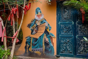 A mural of a monkey dressed in elaborate traditional Chinese attire with intricate designs, set against a mustard-colored wall. The monkey is seated, holding a scroll, and surrounded by a doorway with ornate carvings. Red ribbons hang from nearby trees, adding a splash of color.