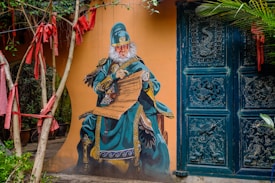 A mural of a monkey dressed in elaborate traditional Chinese attire with intricate designs, set against a mustard-colored wall. The monkey is seated, holding a scroll, and surrounded by a doorway with ornate carvings. Red ribbons hang from nearby trees, adding a splash of color.