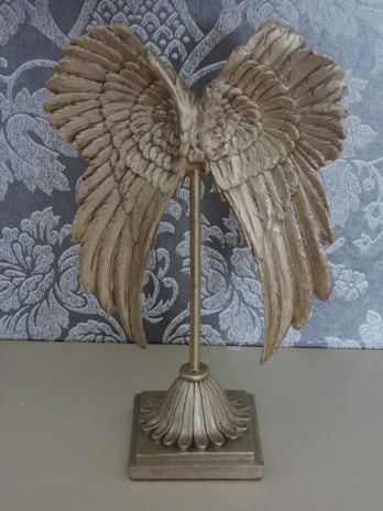 A decorative object resembling golden wings attached to a vertical stand, set against a patterned dark gray and white background.