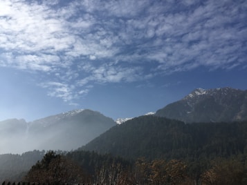 A picturesque landscape featuring towering mountains with snow-capped peaks set against a clear blue sky with scattered clouds. Dense forests cover the lower slopes, and a gentle mist enhances the serene ambiance.