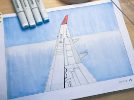 A hand-drawn illustration depicts the view of an airplane wing, labeled with 'B-1758', against a blue sky with clouds. The drawing is surrounded by art supplies including four blue-tinted markers. The paper is dated and signed in the bottom right corner.