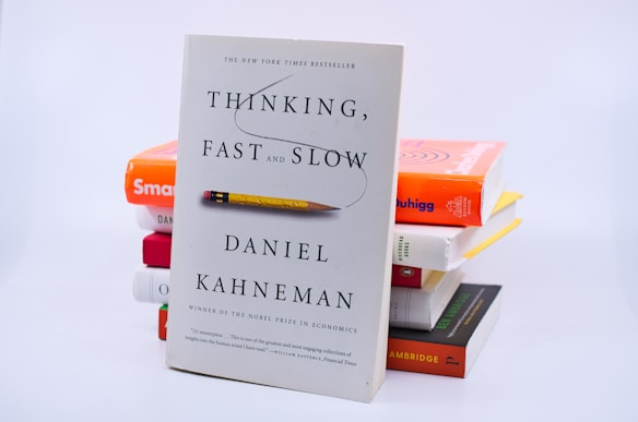 A stack of books with 'Thinking, Fast and Slow' by Daniel Kahneman prominently displayed in the foreground. The cover features a simple design with a pencil graphic. Other books in the stack include brightly colored covers adding a pop of color to the scene.