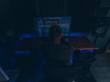 A person is sitting in a dimly lit room in front of a large computer monitor displaying audio editing software. There are two large speakers on either side of the desk, and the room has a dark, cozy atmosphere with blue accent lighting.