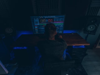 A person is sitting in a dimly lit room in front of a large computer monitor displaying audio editing software. There are two large speakers on either side of the desk, and the room has a dark, cozy atmosphere with blue accent lighting.