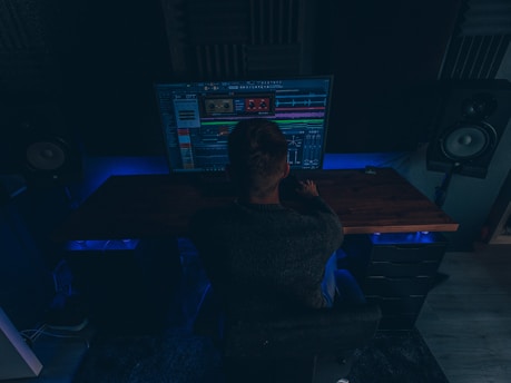 A person is sitting in a dimly lit room in front of a large computer monitor displaying audio editing software. There are two large speakers on either side of the desk, and the room has a dark, cozy atmosphere with blue accent lighting.
