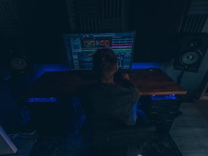 A person is sitting in a dimly lit room in front of a large computer monitor displaying audio editing software. There are two large speakers on either side of the desk, and the room has a dark, cozy atmosphere with blue accent lighting.