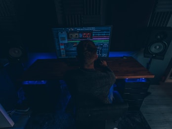 A person is sitting in a dimly lit room in front of a large computer monitor displaying audio editing software. There are two large speakers on either side of the desk, and the room has a dark, cozy atmosphere with blue accent lighting.