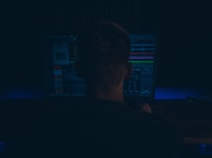 A person is seated in front of a computer monitor displaying software with various waveforms and graphs, suggesting audio editing or production work. The setting is dimly lit, creating a focused and immersive atmosphere.