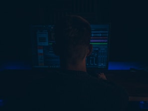 A person is seated in front of a computer monitor displaying software with various waveforms and graphs, suggesting audio editing or production work. The setting is dimly lit, creating a focused and immersive atmosphere.