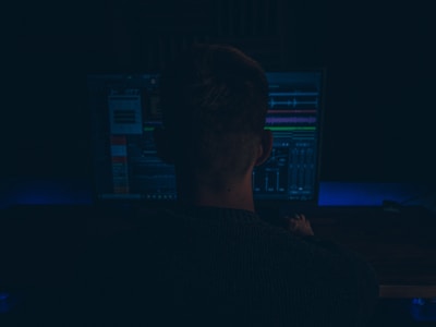 A person is seated in front of a computer monitor displaying software with various waveforms and graphs, suggesting audio editing or production work. The setting is dimly lit, creating a focused and immersive atmosphere.