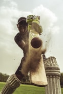 A cricket player in full gear is captured hitting a cricket ball with a bat. The image focuses on the moment of impact as dust and particles scatter from the bat. The player is wearing pads, gloves, and a helmet, and the background shows a green field and a cloudy sky.