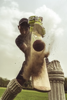 A cricket player in full gear is captured hitting a cricket ball with a bat. The image focuses on the moment of impact as dust and particles scatter from the bat. The player is wearing pads, gloves, and a helmet, and the background shows a green field and a cloudy sky.