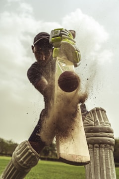 A cricket player in full gear is captured hitting a cricket ball with a bat. The image focuses on the moment of impact as dust and particles scatter from the bat. The player is wearing pads, gloves, and a helmet, and the background shows a green field and a cloudy sky.