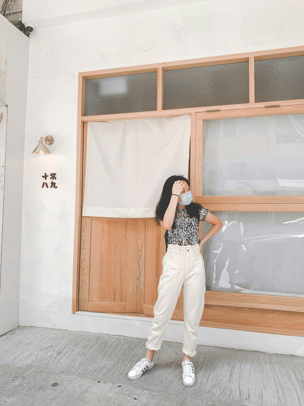 mulher na camisa de bolinhas preta e branca e calças brancas de pé ao lado da porta de madeira marrom