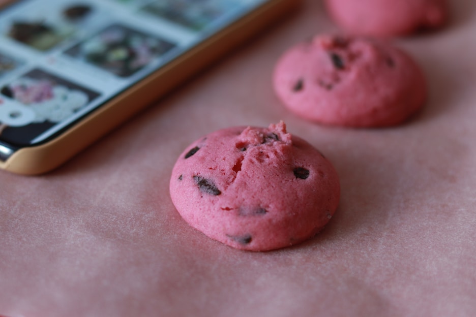 Pink cookies with dark chocolate chips are placed on a pink surface next to a smartphone displaying images of similar cookies.