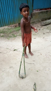 A young child stands on a sandy surface holding a handmade bamboo toy with wheels. The child is barefoot, wearing a red patterned cloth and a necklace. Behind is a blue corrugated metal wall and some greenery.
