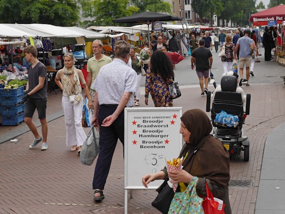 A busy outdoor market scene with people walking and browsing stalls. Various goods like fresh produce and flowers are on display. A sign in the foreground advertises food items in Dutch. The atmosphere is lively with shoppers carrying bags and interacting. A woman in a brown headscarf is seen in the front holding fries.