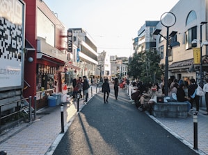 A busy urban street lined with shops and cafés, people walking and sitting along the sides. Brightly colored signs and advertisements adorn the buildings, and a large QR code is visible on the left side. The sunlight casts long shadows on the pavement, suggesting it's late afternoon.