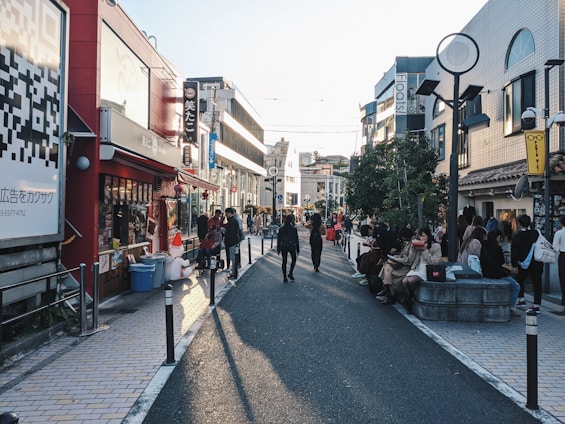 A busy urban street lined with shops and cafés, people walking and sitting along the sides. Brightly colored signs and advertisements adorn the buildings, and a large QR code is visible on the left side. The sunlight casts long shadows on the pavement, suggesting it's late afternoon.