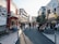 A busy urban street lined with shops and cafés, people walking and sitting along the sides. Brightly colored signs and advertisements adorn the buildings, and a large QR code is visible on the left side. The sunlight casts long shadows on the pavement, suggesting it's late afternoon.