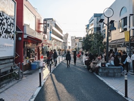 A busy urban street lined with shops and caf&eacute;s, people walking and sitting along the sides. Brightly colored signs and advertisements adorn the buildings, and a large QR code is visible on the left side. The sunlight casts long shadows on the pavement, suggesting it's late afternoon.