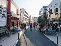 A busy urban street lined with shops and caf&eacute;s, people walking and sitting along the sides. Brightly colored signs and advertisements adorn the buildings, and a large QR code is visible on the left side. The sunlight casts long shadows on the pavement, suggesting it's late afternoon.