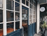 A cozy cafe with large windows and a blue door, featuring a person inside wearing a patterned hat. The cafe sign reads 'Onigily Cafe' with a logo of cutlery. A building with an urban landscape is visible in the reflection of the glass, along with a plant near the entrance.