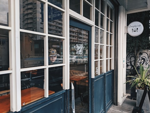A cozy cafe with large windows and a blue door, featuring a person inside wearing a patterned hat. The cafe sign reads 'Onigily Cafe' with a logo of cutlery. A building with an urban landscape is visible in the reflection of the glass, along with a plant near the entrance.