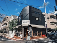 A modern, black building with large windows and a sign reading 'MARZAC 7' is situated on a street corner. The building has an awning and a small outdoor seating area with wooden accents. It is surrounded by urban residential buildings and utility poles with a clear, blue sky overhead.