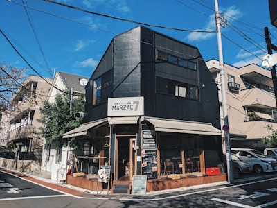 A modern, black building with large windows and a sign reading 'MARZAC 7' is situated on a street corner. The building has an awning and a small outdoor seating area with wooden accents. It is surrounded by urban residential buildings and utility poles with a clear, blue sky overhead.