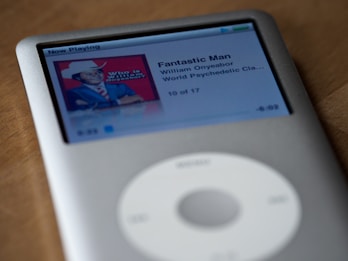 A silver iPod Classic displaying a music track on its screen. The track is titled 'Fantastic Man' by William Onyeabor from the album 'World Psychedelic Classics'. The album art features a person in a suit and hat against a red background.