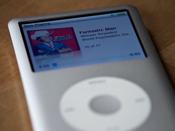 A silver iPod Classic displaying a music track on its screen. The track is titled 'Fantastic Man' by William Onyeabor from the album 'World Psychedelic Classics'. The album art features a person in a suit and hat against a red background.