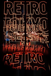 Bright neon sign featuring the words 'RETRO TOKYO TECH' in orange-red hues, with a reflection creating a mirrored effect. Below the neon, several sneakers are displayed.