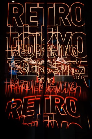 Bright neon sign featuring the words 'RETRO TOKYO TECH' in orange-red hues, with a reflection creating a mirrored effect. Below the neon, several sneakers are displayed.